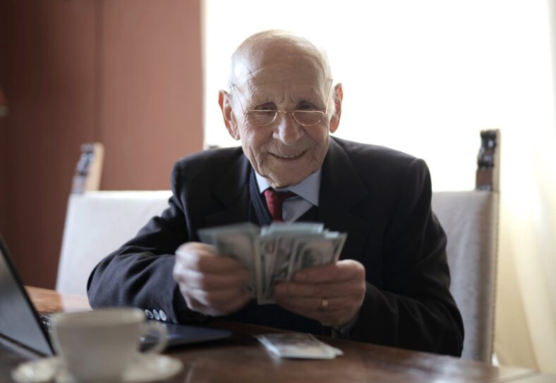 happy senior businessman counting money while sitting at table with laptop
